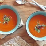 Two bowls of vibrant tomato soup garnished with fresh microgreens on a wooden table.