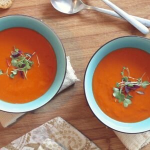 Two bowls of vibrant tomato soup garnished with fresh microgreens on a wooden table.