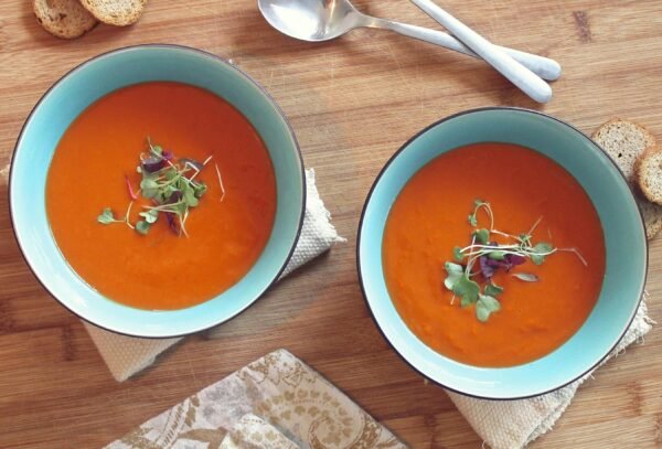 Two bowls of vibrant tomato soup garnished with fresh microgreens on a wooden table.